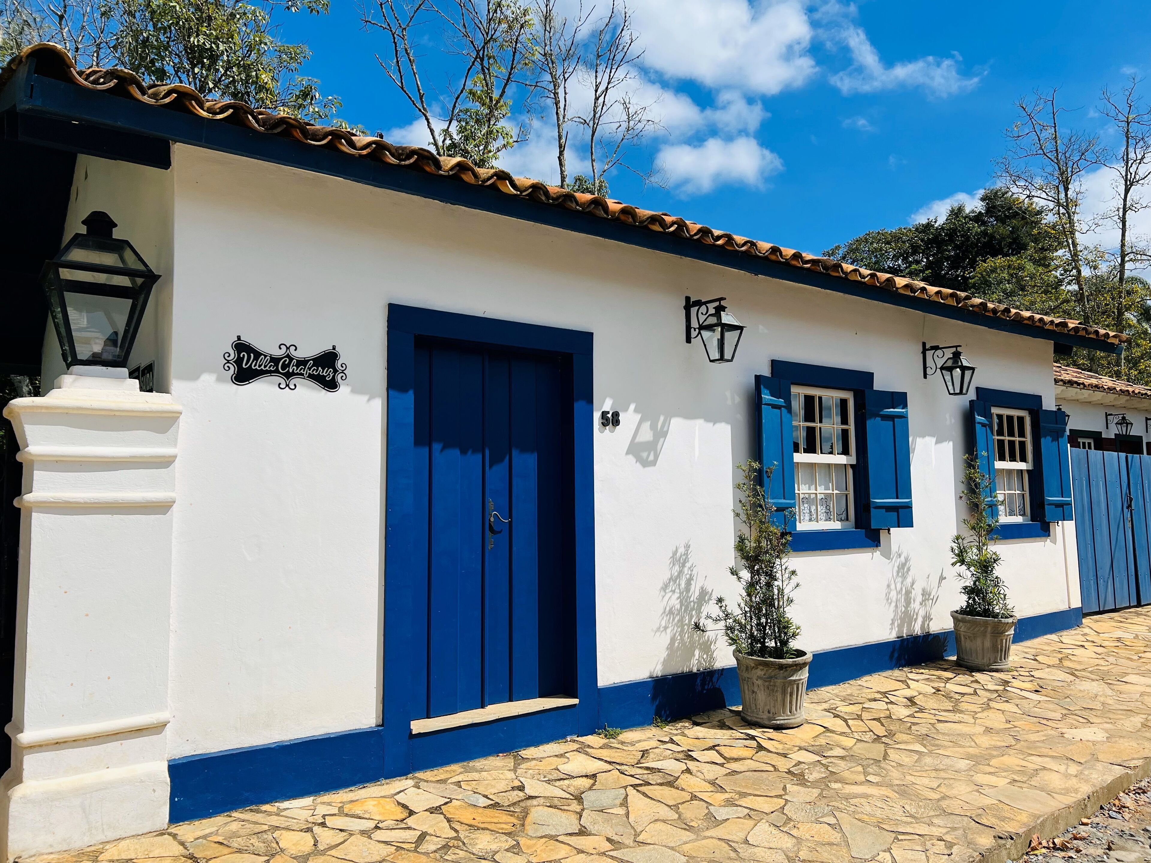 A white building with blue doors and shutters, a stone-paved entrance, and a sign reading "Villa Chafariz".