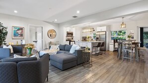 A spacious living area with a grey sofa, a coffee table, and a dining area with bar stools. - Hot Tub, Firepit, Deck | 10mins to Beach | Middle Terrace Cottage by AvantStay (Fort Lauderdale)