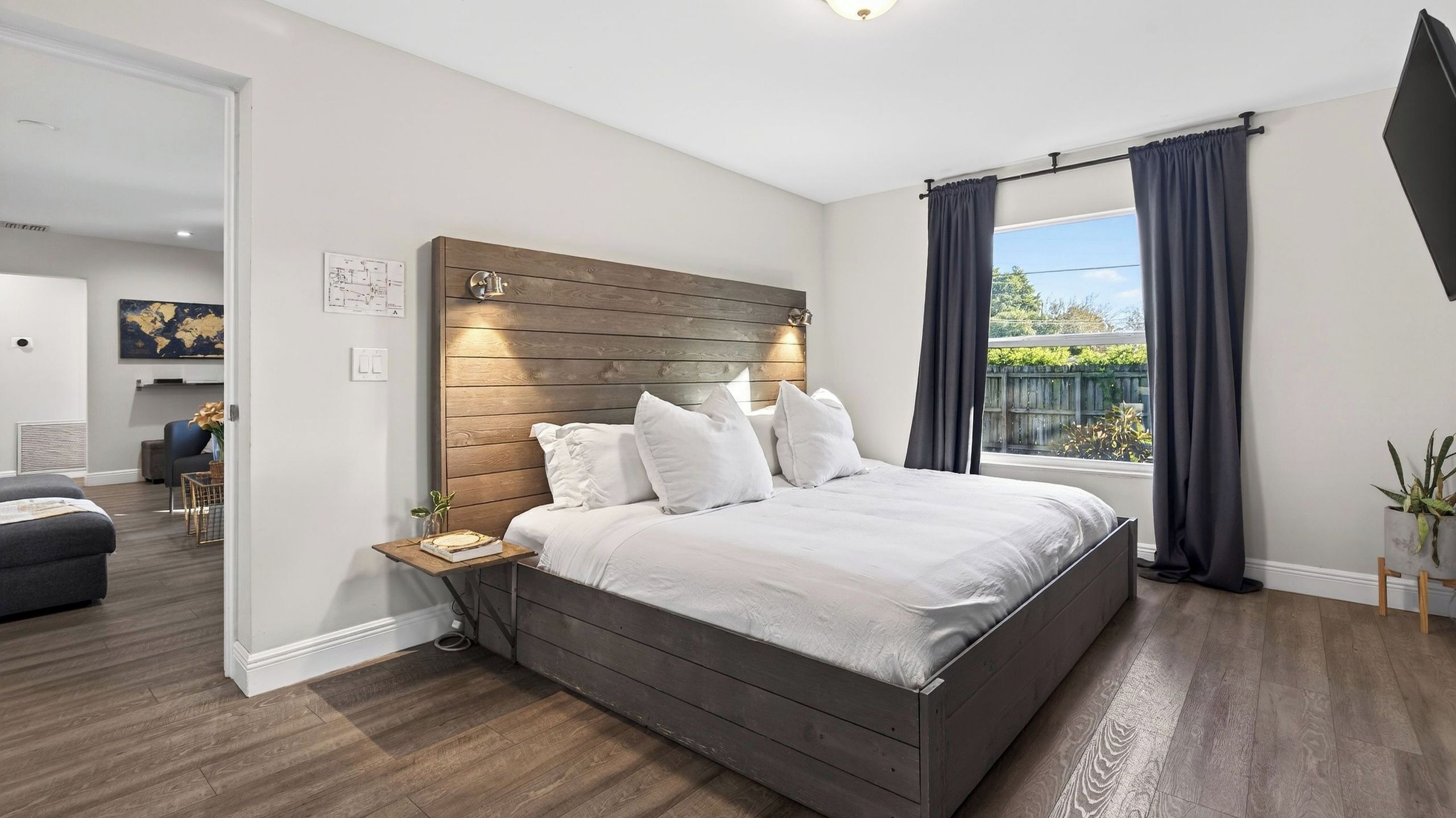 Une chambre moderne avec une tête de lit en bois, un grand lit, une table de chevet, une fenêtre avec des rideaux et une vue sur l’extérieur.
