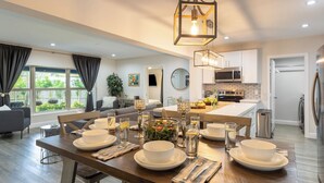 A modern kitchen with white cabinets, stainless steel appliances, and a dining area with a wooden table set for four. - Hot Tub, Firepit, Deck | 10mins to Beach | Middle Terrace Cottage by AvantStay (Fort Lauderdale)