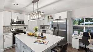 A modern kitchen with white cabinetry, stainless steel appliances, and a central island with a magazine and decorative items. - Hot Tub, Firepit, Deck | 10mins to Beach | Middle Terrace Cottage by AvantStay (Fort Lauderdale)