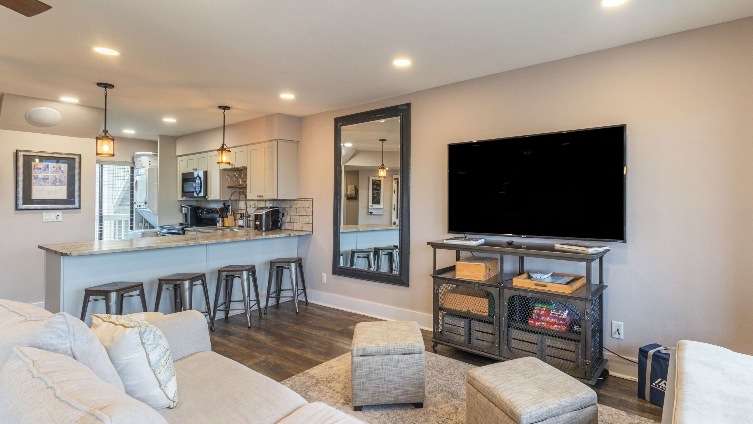 A modern living room with a large TV, a kitchen area with bar stools, and a dining area with a large mirror.