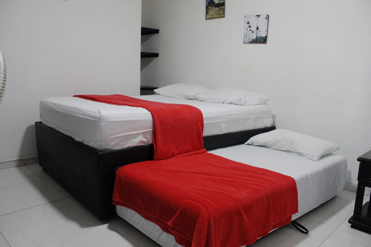 Quarto com duas camas, cada uma com roupa de cama branca e um cobertor vermelho. Há uma pequena mesa e uma cadeira no canto.