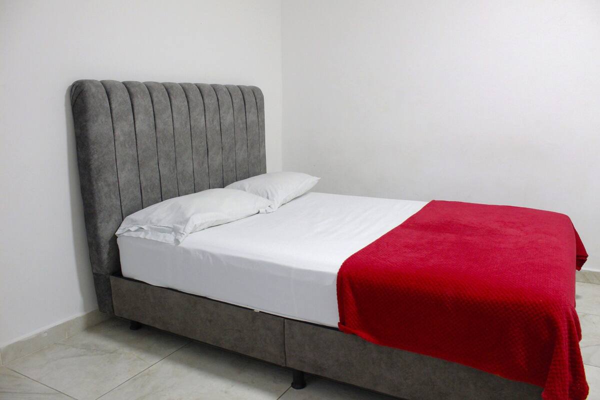 Uma cama de solteiro com cabeceira cinza revestida em tecido e roupa de cama branca, um cobertor vermelho e uma almofada branca.