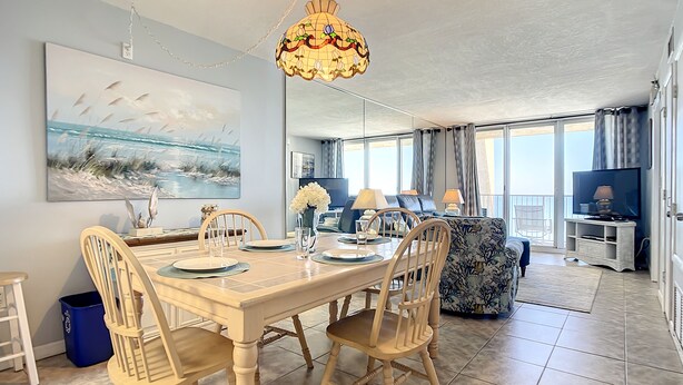 A dining area with a wooden table set for four, surrounded by chairs. There is a large painting on the wall, a decorative hanging light, and a television in the background. - Direct Oceanfront Condo - Pool, Beach & Steps from Flagler Ave (New Smyrna Beach)