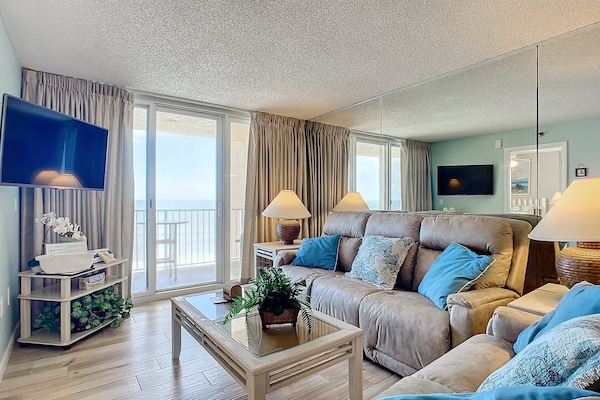 Relax in a plush, ocean-inspired living room featuring mirrored walls, a balcony view, and a mounted TV for ultimate comfort and entertainment.