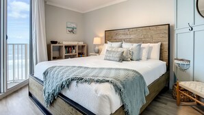 A bedroom with a wooden headboard, a large bed with multiple pillows, a nightstand with a lamp, a bookshelf, and a view of the ocean through a sliding door. - Impressive Oceanfront Condo - Pool, BBQ & Just Steps to Flagler Ave (New Smyrna Beach)