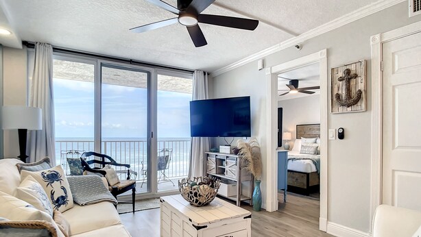A spacious living room with a white sofa, a TV, and a balcony with a view of the ocean. - Impressive Oceanfront Condo - Pool, BBQ & Just Steps to Flagler Ave (New Smyrna Beach)