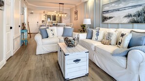 A spacious living room with a white sofa set, a wooden coffee table, and a ceiling fan. - Impressive Oceanfront Condo - Pool, BBQ & Just Steps to Flagler Ave (New Smyrna Beach)