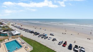 Blick auf einen Strand mit Swimmingpool, Strandsonnenschirmen und einem Parkplatz.
