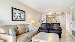 A living room with a beige sofa, a dark blue sofa, and a wooden coffee table. There is a painting on the wall and a lamp on the side table.