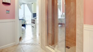 A hallway leading to a bathroom with a glass shower enclosure, a small seating area with a TV, and a balcony visible through the door.