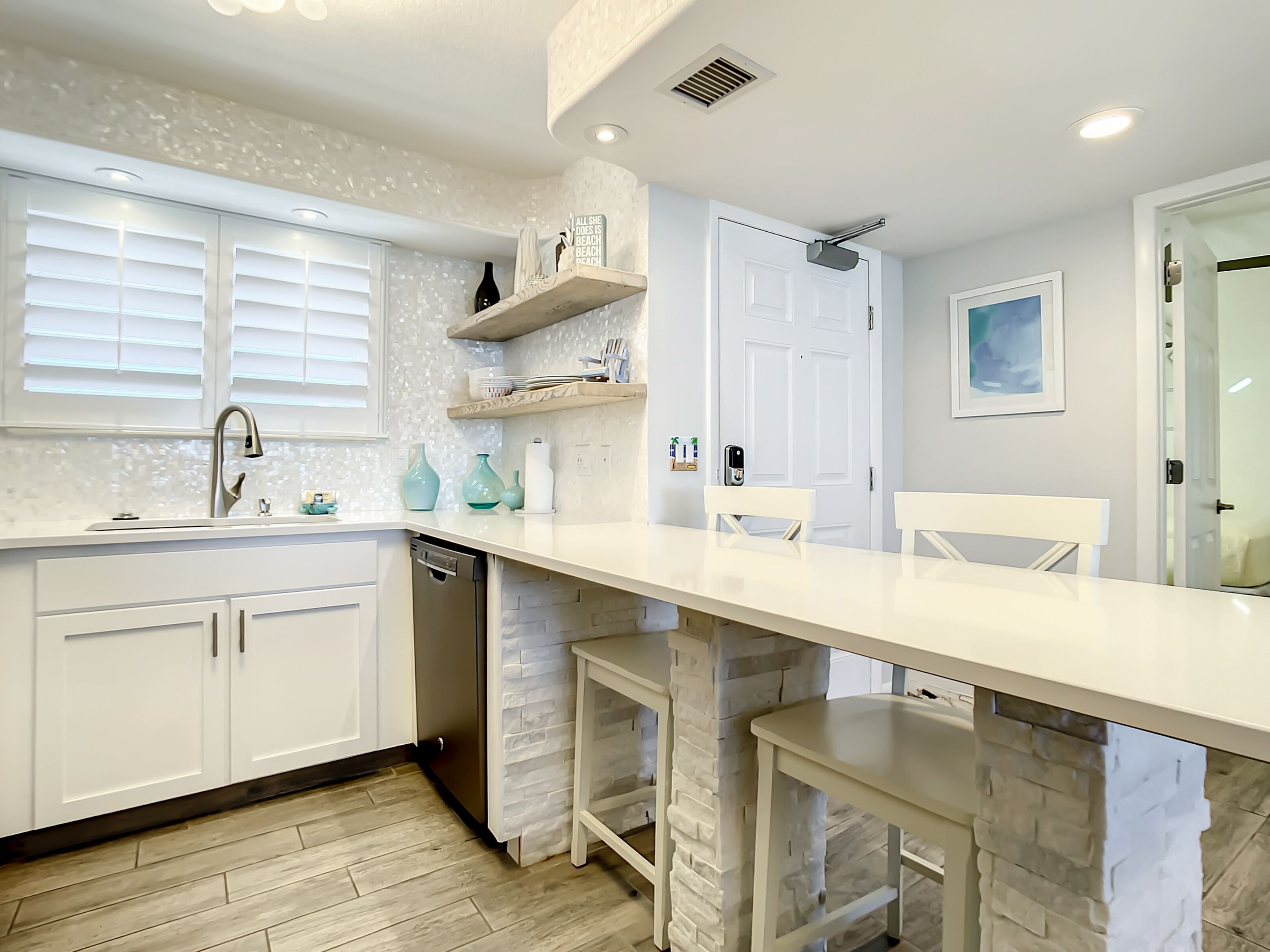 A modern kitchen with white cabinets, a white countertop, and a stone accent wall. There is a sink with a faucet, a dish rack, and a built-in oven.