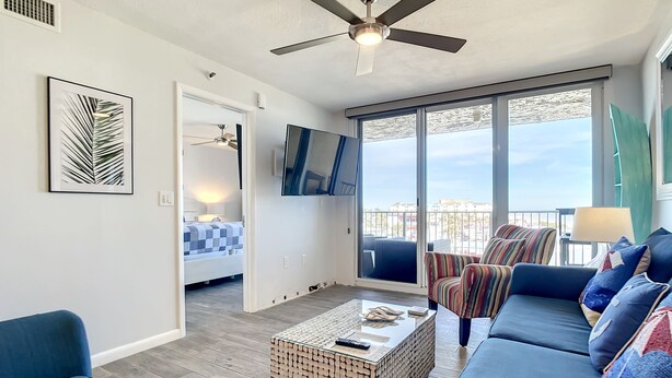 Un salon moderne avec un canapé bleu, une table basse et un ventilateur de plafond. Il y a un balcon avec vue sur la ville.
