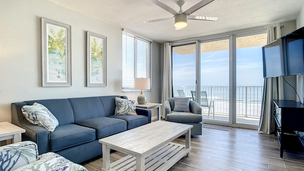 A spacious living area with a blue sofa, a white coffee table, and a grey armchair. There are two framed pictures on the wall, a ceiling fan, and a large window offering a view of the beach.