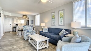 A spacious living room with a blue sofa, a white coffee table, and a patterned armchair. There is a ceiling fan, a kitchen in the background, and a dining area with a white table and chairs.