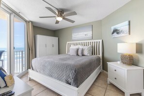 Une chambre à coucher avec un lit, une table de chevet, un ventilateur de plafond, une fenêtre avec des rideaux et une vue sur l’océan.
