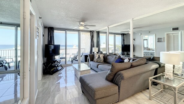 A spacious living room with a large sofa, a glass coffee table, and a flat-screen TV. The room has a balcony with a view of the ocean. - Bright Oceanview Condo • Steps to Sand & Flagler Dining Scene (New Smyrna Beach)