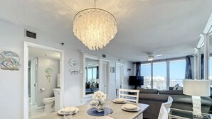 A modern living room with a dining area, a large chandelier, and a view of the city. - Bright Oceanview Condo • Steps to Sand & Flagler Dining Scene (New Smyrna Beach)