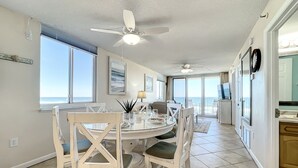 A spacious dining area with a round table, white chairs, and a view of the ocean through large windows. - Beachfront, Pool, BBQ & Game room!  Extra Windows = More Light & More Views (New Smyrna Beach)