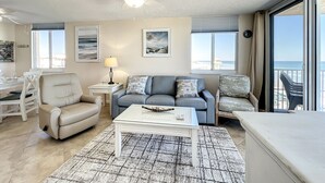 A spacious living room with a ceiling fan, a sofa set, a coffee table, and a view of the ocean through large windows. - Beachfront, Pool, BBQ & Game room!  Extra Windows = More Light & More Views (New Smyrna Beach)