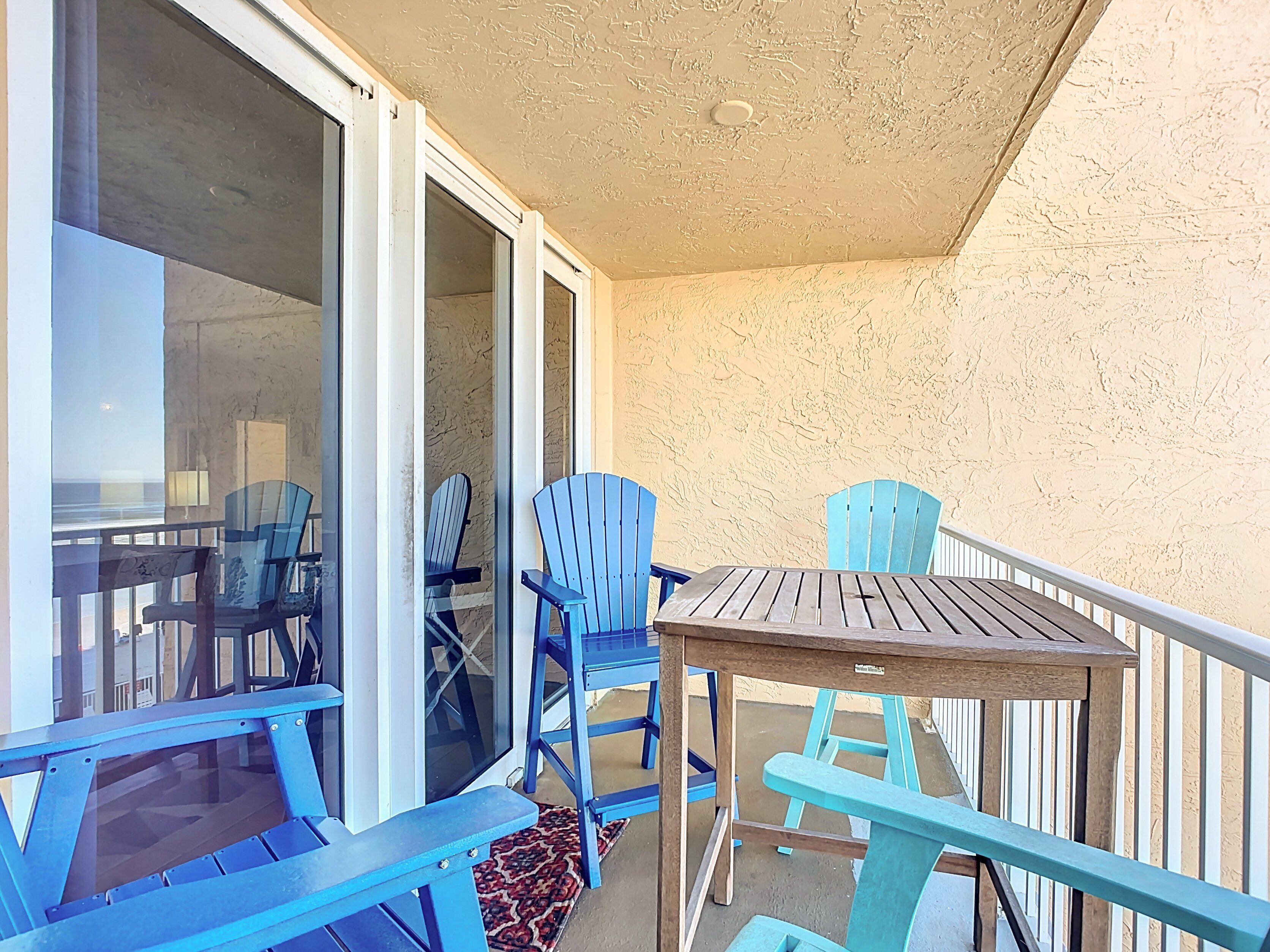 Ein Balkon mit blauen Stühlen und einem Holztisch, einer Schiebetür aus Glas und einer strukturierten Wand.