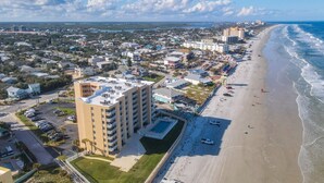 An aerial view of a coastal city with a prominent hotel featuring multiple floors, a parking lot, and a beachfront. - Updated 2BR oceanfront condo with balcony & pool (New Smyrna Beach)