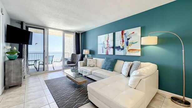A modern living room with a white sofa, a glass coffee table, and a teal accent wall. - Updated 2BR beachfront condo with balcony + pool (New Smyrna Beach)