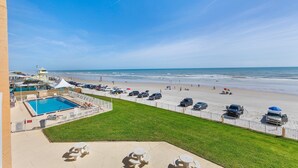 A beach view from a hotel balcony with a swimming pool, beach chairs, and a fenced parking area. - Updated Oceanfront Condo!  Pool, Beach & steps from Flagler Ave (New Smyrna Beach)
