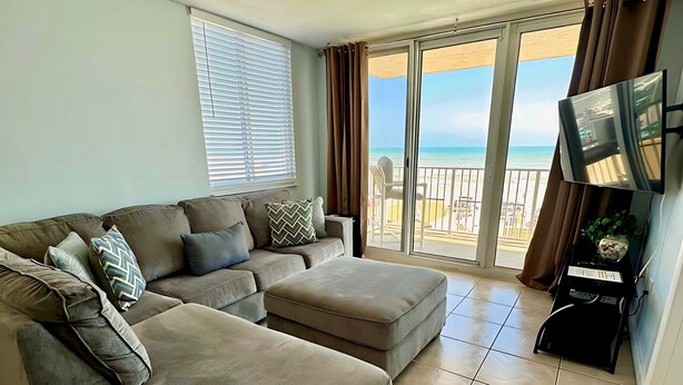 A living room with a sofa, ottoman, and a TV on a wall. - OceanFront Condo - Pool, BBQ, Steps from Flagler Ave (New Smyrna Beach)