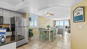 A modern kitchen with a stainless steel refrigerator, a dining area with green chairs, and a ceiling fan. - OceanFront Condo - Pool, BBQ, Steps from Flagler Ave (New Smyrna Beach)