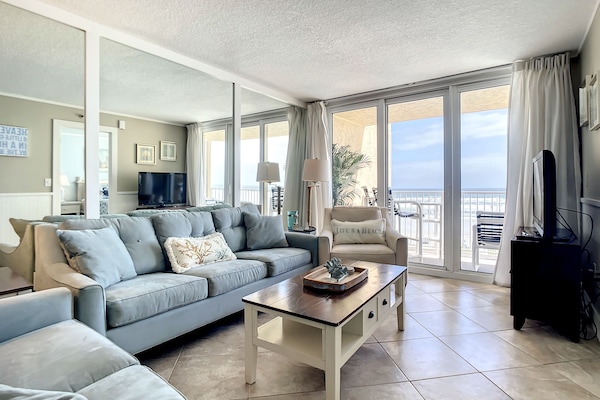 This airy living room features plush seating, a coffee table, and expansive windows offering stunning ocean views. The comfortable and inviting atmosphere is perfect for relaxing or watching TV after a day at the beach.