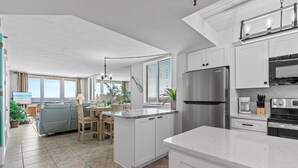 A modern kitchen with white cabinetry, a stainless steel refrigerator, and a dining area with a view of the sea.
