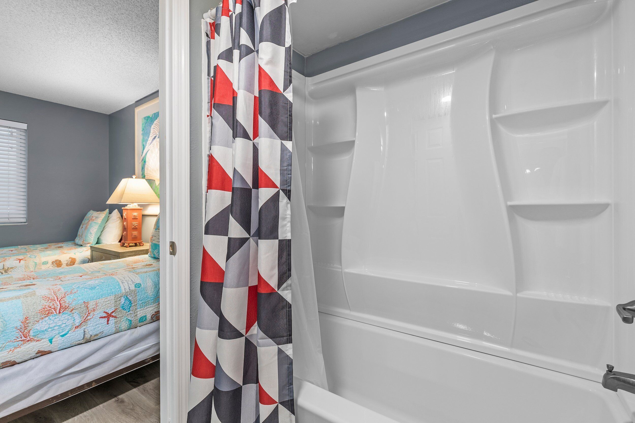 A bathroom with a white bathtub, a shower curtain with a geometric pattern, and a bed with a colorful bedspread.