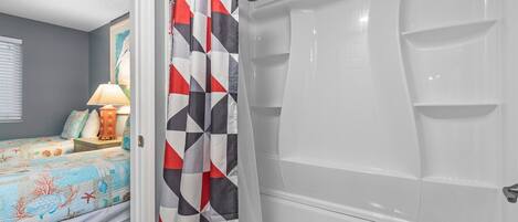 A bathroom with a white bathtub, a shower curtain with a geometric pattern, and a bed with a colorful bedspread.