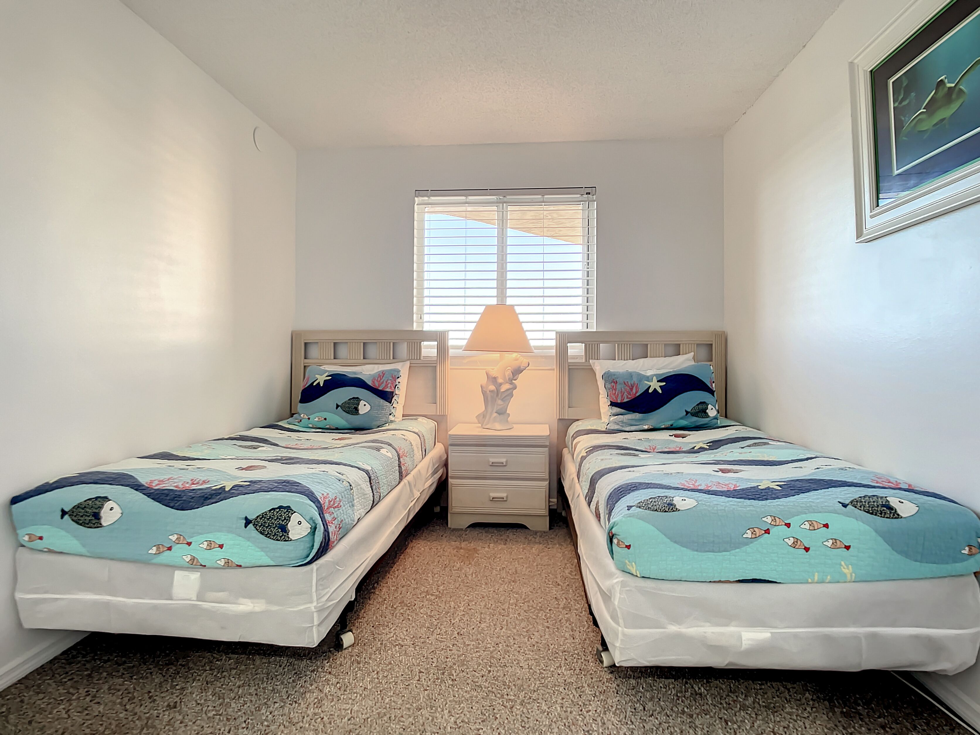 A bedroom with two single beds, a nightstand with a lamp, a window with blinds, and a framed picture on the wall.