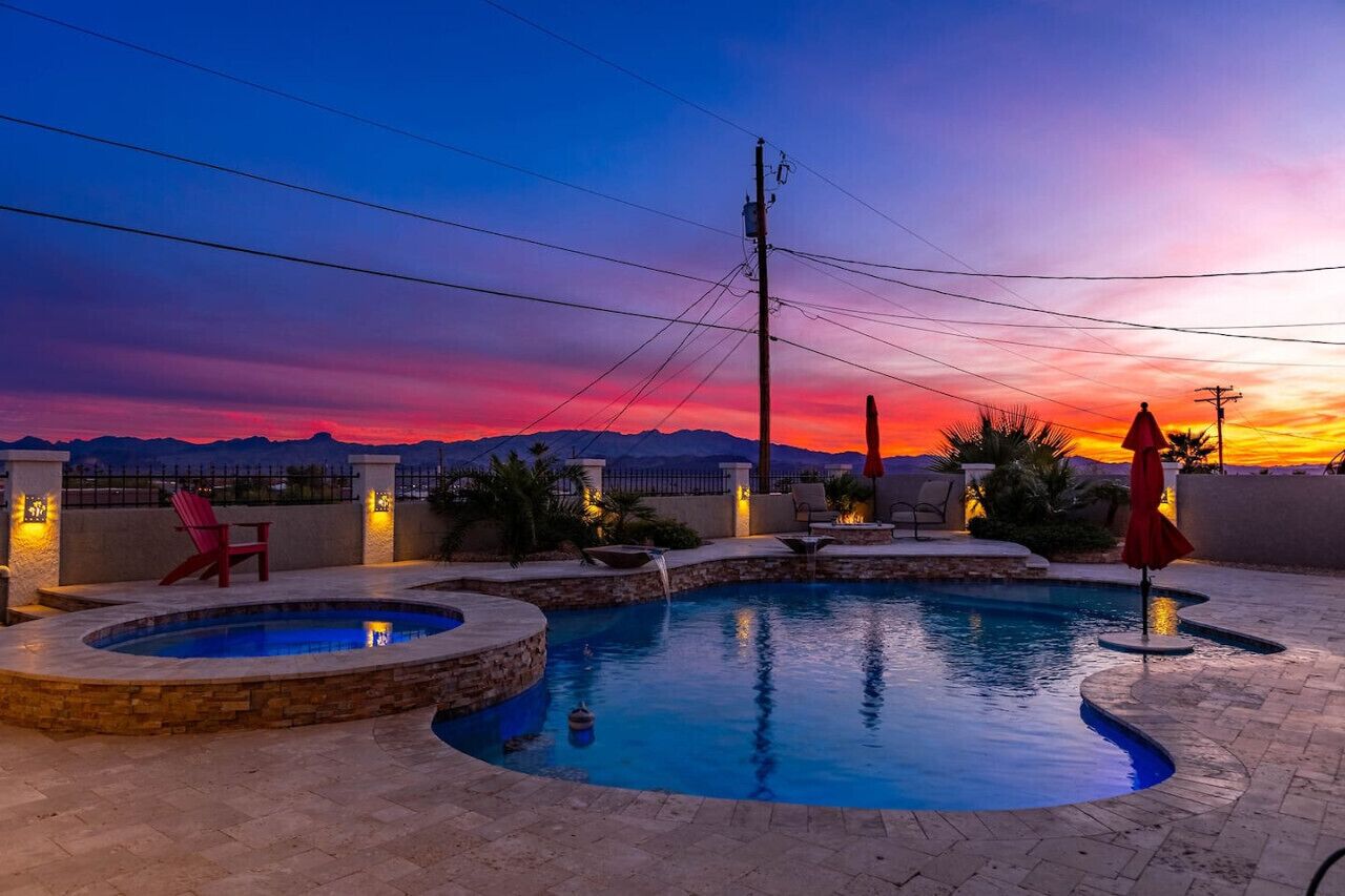 Une piscine avec un jacuzzi, entourée de chaises longues et de parasols, le tout mis en valeur par un coucher de soleil et un paysage montagneux en arrière-plan.