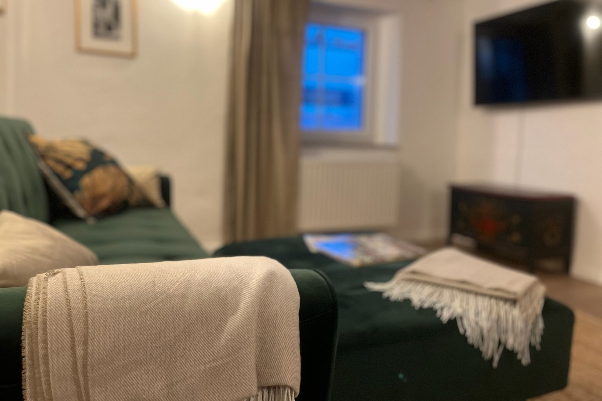 Olohuoneessa on vihreä sohva, beigenvärinen heittopeitto ja seinälle asennettu televisio.