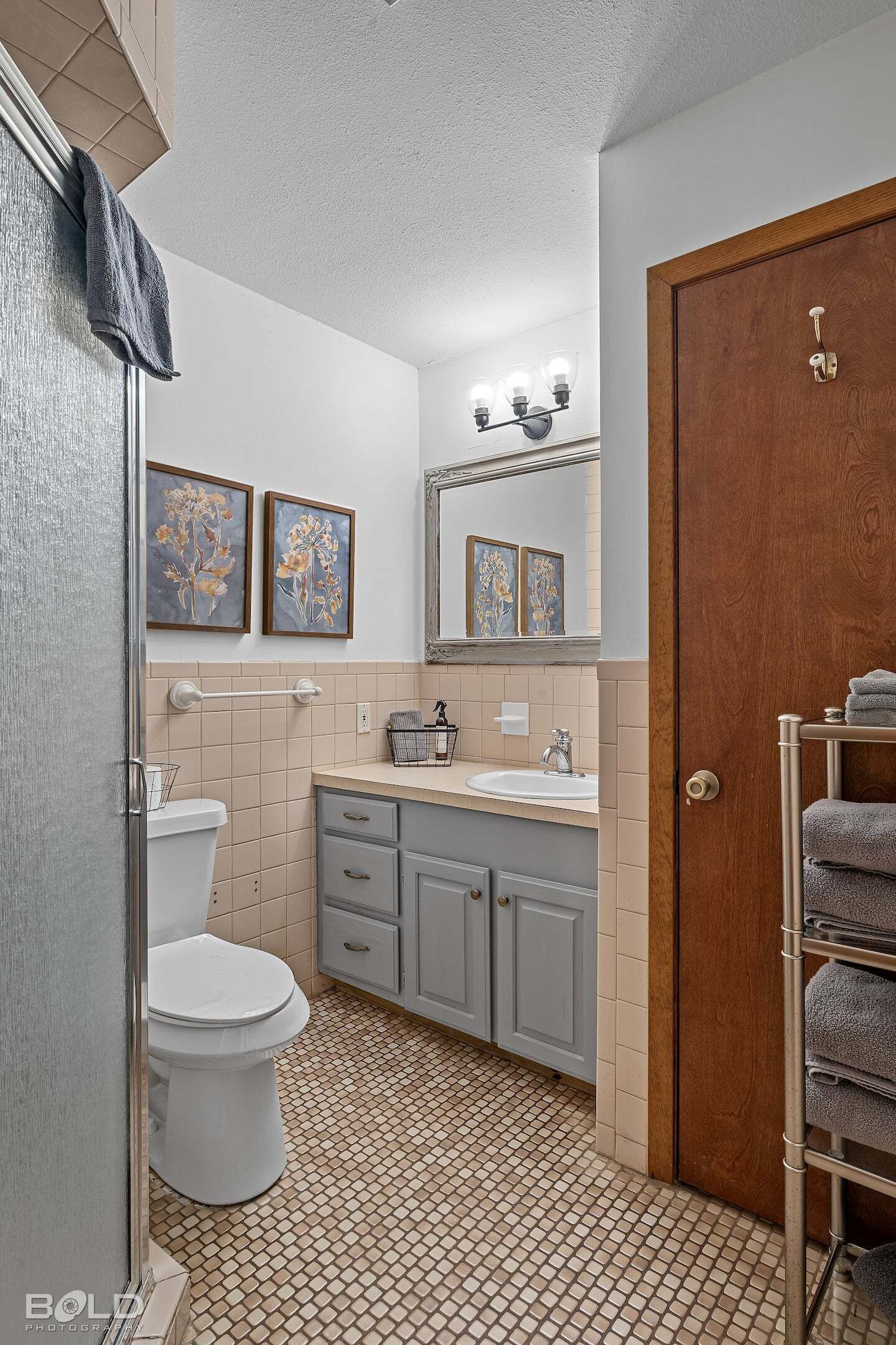 Een badkamer met toilet, wastafel en wastafelmeubel. Er hangen twee geborduurde bloemschilderijen aan de muur, een handdoekhouder en een houten deur.