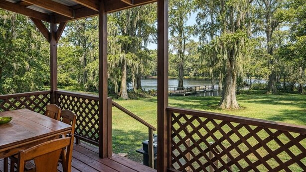 A covered outdoor area with a wooden table and chairs, a lattice railing, and a view of a lake surrounded by trees. - Lakefront cabin with sweeping views of Caddo Lake, lakeside to the pier and kayaks, plus a foosball table for fun downtime. (Karnack)