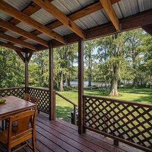 A covered outdoor area with a wooden table and chairs, a lattice railing, and a view of a lake surrounded by trees. - Lakefront cabin with sweeping views of Caddo Lake, lakeside to the pier and kayaks, plus a foosball table for fun downtime. (Karnack)