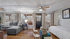 A spacious room with a gray sofa, a wooden dining table with chairs, a bed, and a ceiling fan. - Lakefront cabin with sweeping views of Caddo Lake, lakeside to the pier and kayaks, plus a foosball table for fun downtime. (Karnack)