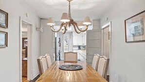 A dining room with a wooden table, six chairs, a chandelier, and a view into the kitchen. - Lakefront cabin with sweeping views of Caddo Lake, lakeside to the pier and kayaks, plus a foosball table for fun downtime. (Karnack)