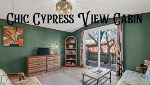 A cozy cabin interior with a television, bookshelf, and a view of the outdoors. - Lakefront cabin with sweeping views of Caddo Lake, lakeside to the pier and kayaks, plus a foosball table for fun downtime. (Karnack)