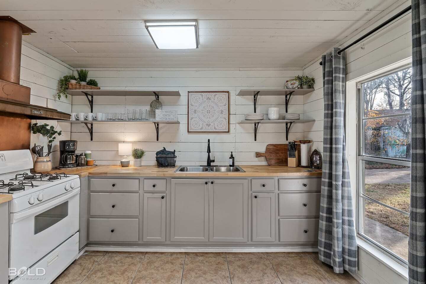 Une cuisine avec des meubles blancs, un plan de travail en bois, une cuisinière et une fenêtre avec des rideaux.