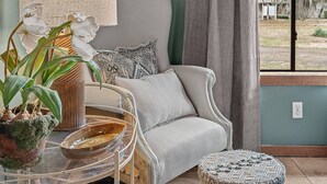 Une pièce avec un mur de couleur vert clair, un fauteuil gris, un pouf rond, une table en verre avec une lampe et une fenêtre donnant sur des arbres.