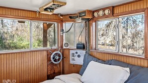 A cabin interior with wooden walls, a bed with gray and white bedding, a steering wheel, and a radio. - Unforgettable Experience: Enjoy a maritime adventure unlike any other. Unwind in a cozy little ship with upper and lower decks with private dock access (Karnack)