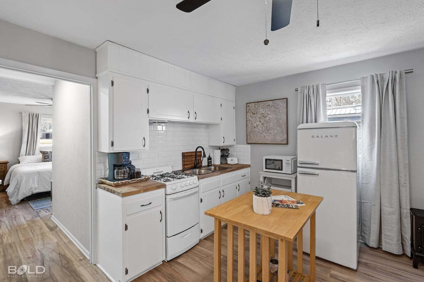 A compact kitchen with white cabinets, a gas stove, a microwave, and a refrigerator. There is a wooden table with a plant and a magazine.
