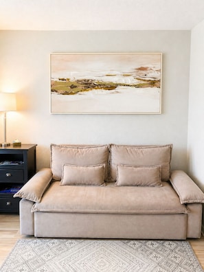 Ein beiges Sofa mit zwei Kissen, ein gemustertes Teppich und ein gerahmtes Landschaftsbild an der Wand.