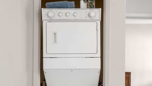 A compact laundry room with a stacked washer and dryer, a towel on the dryer, and a small shelf with two cans.